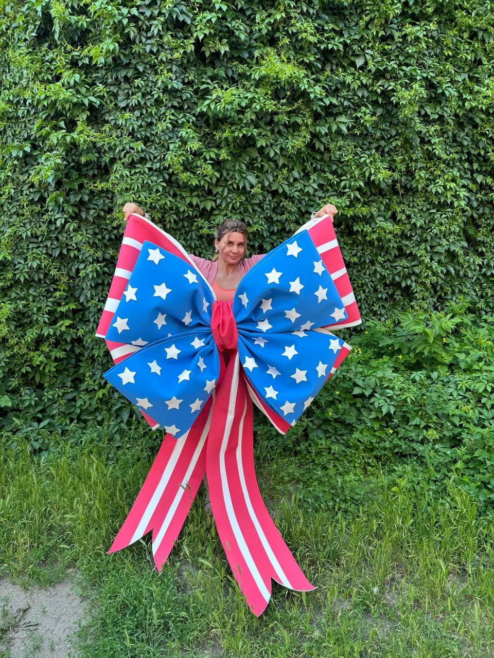 May include: A large decorative bow featuring a patriotic design with red and white stripes and blue sections adorned with white stars. The bow is held up against a backdrop of green foliage and grass.