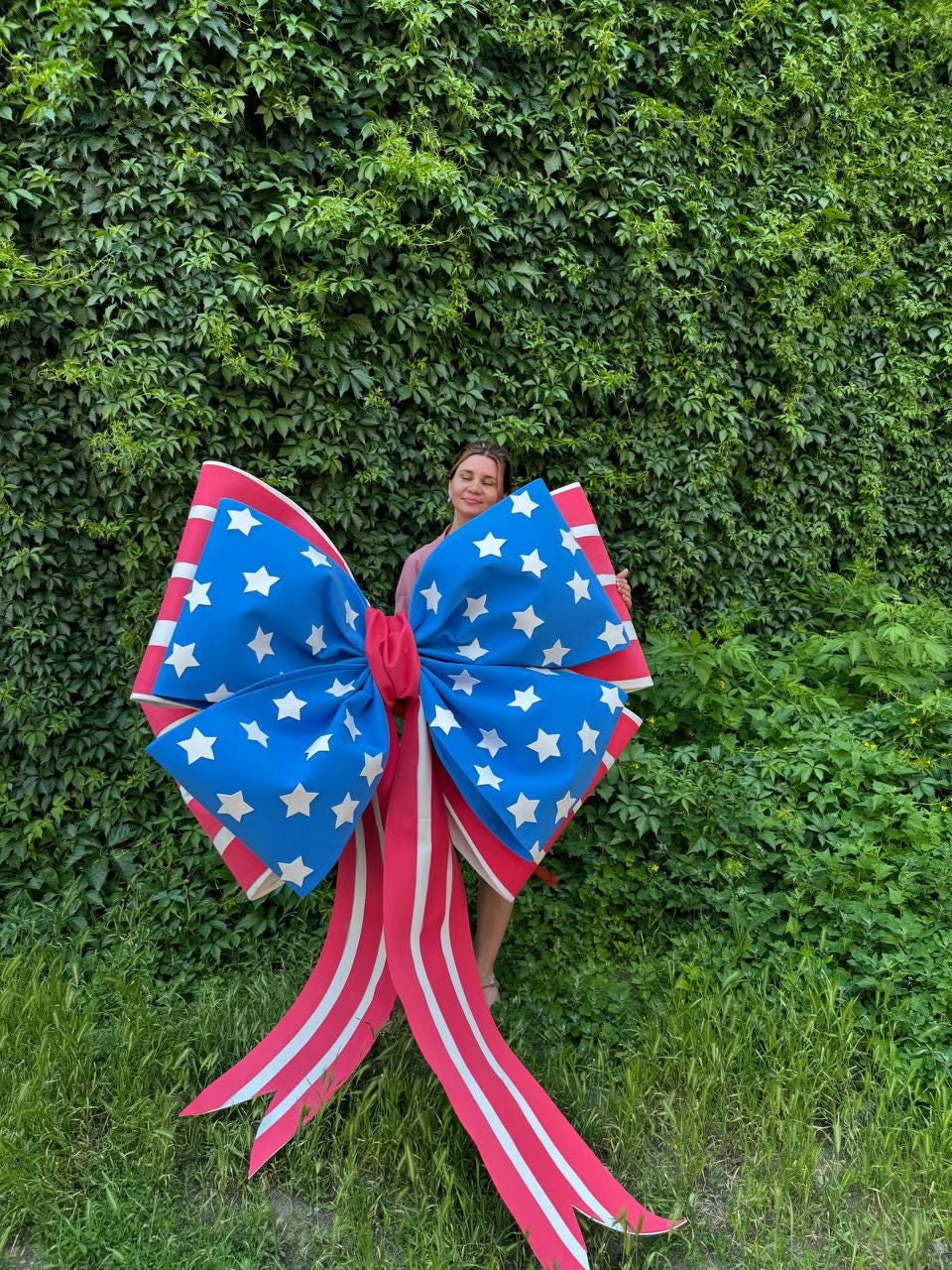 May include: A large decorative bow in patriotic colors. The bow features a blue section with white stars, red edges, and red and white striped streamers. The bow is designed for outdoor decoration.
