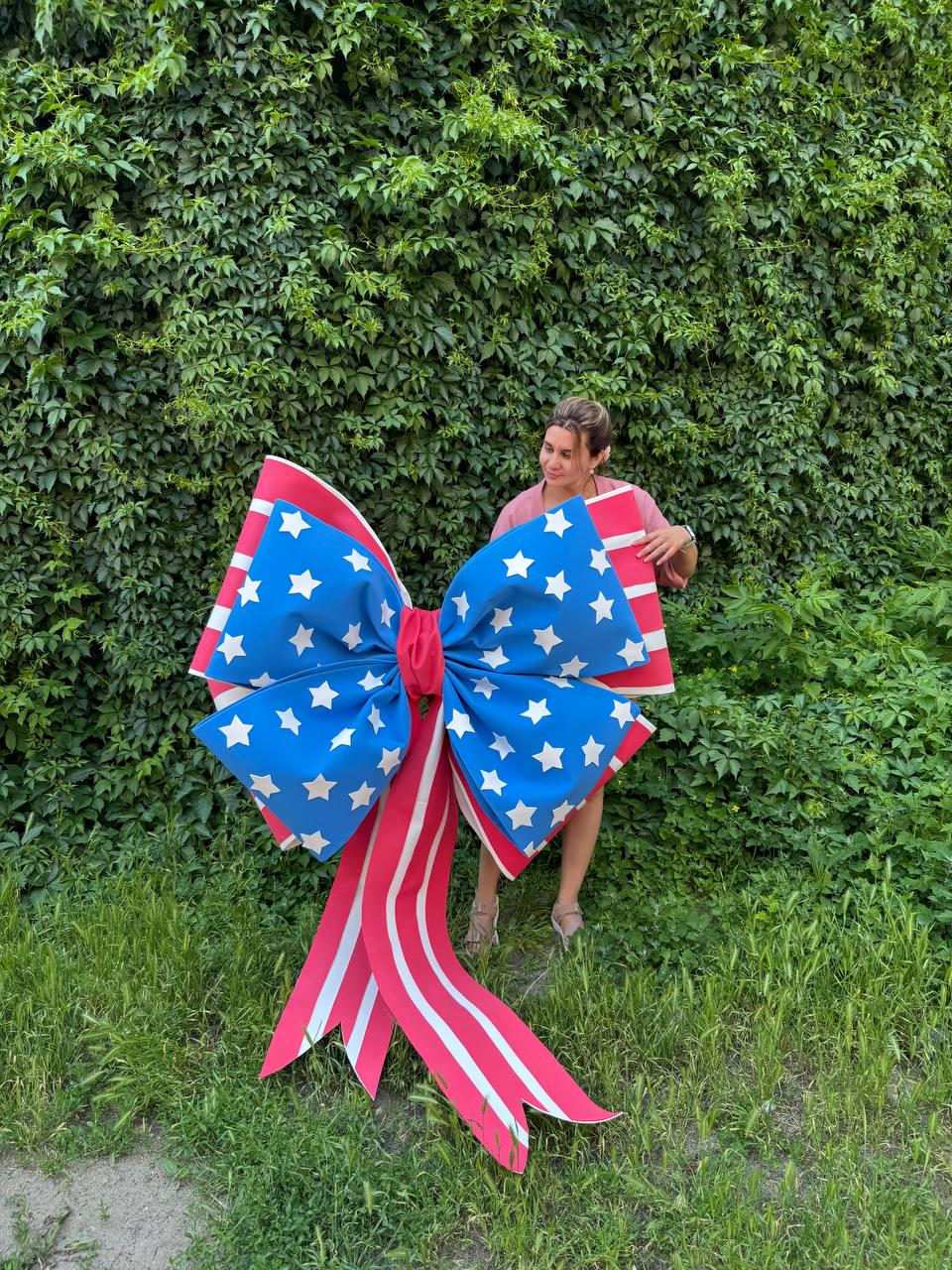 May include: Large patriotic bow decoration with a blue center adorned with white stars and red and white striped edges and tails. The bow is displayed outdoors against a backdrop of green foliage and grass.