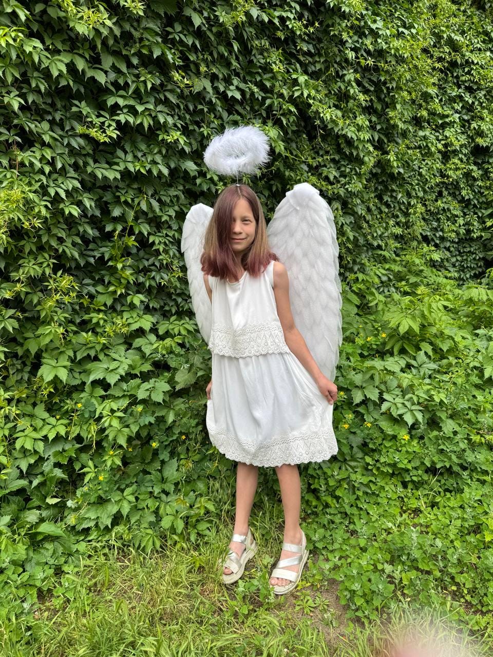 May include: A young person dressed as an angel, wearing a white dress with lace trim, large white feathered wings, and a halo. The background is a lush green wall of foliage, and the person is wearing silver sandals.