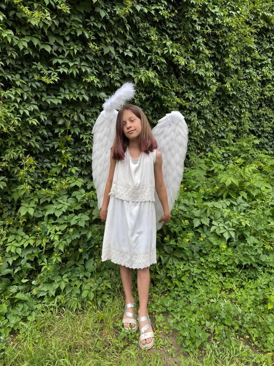 May include: A young person wearing a white dress with lace detailing, silver sandals, and large white angel wings. A white halo sits atop their head. The background is a lush green wall of foliage.