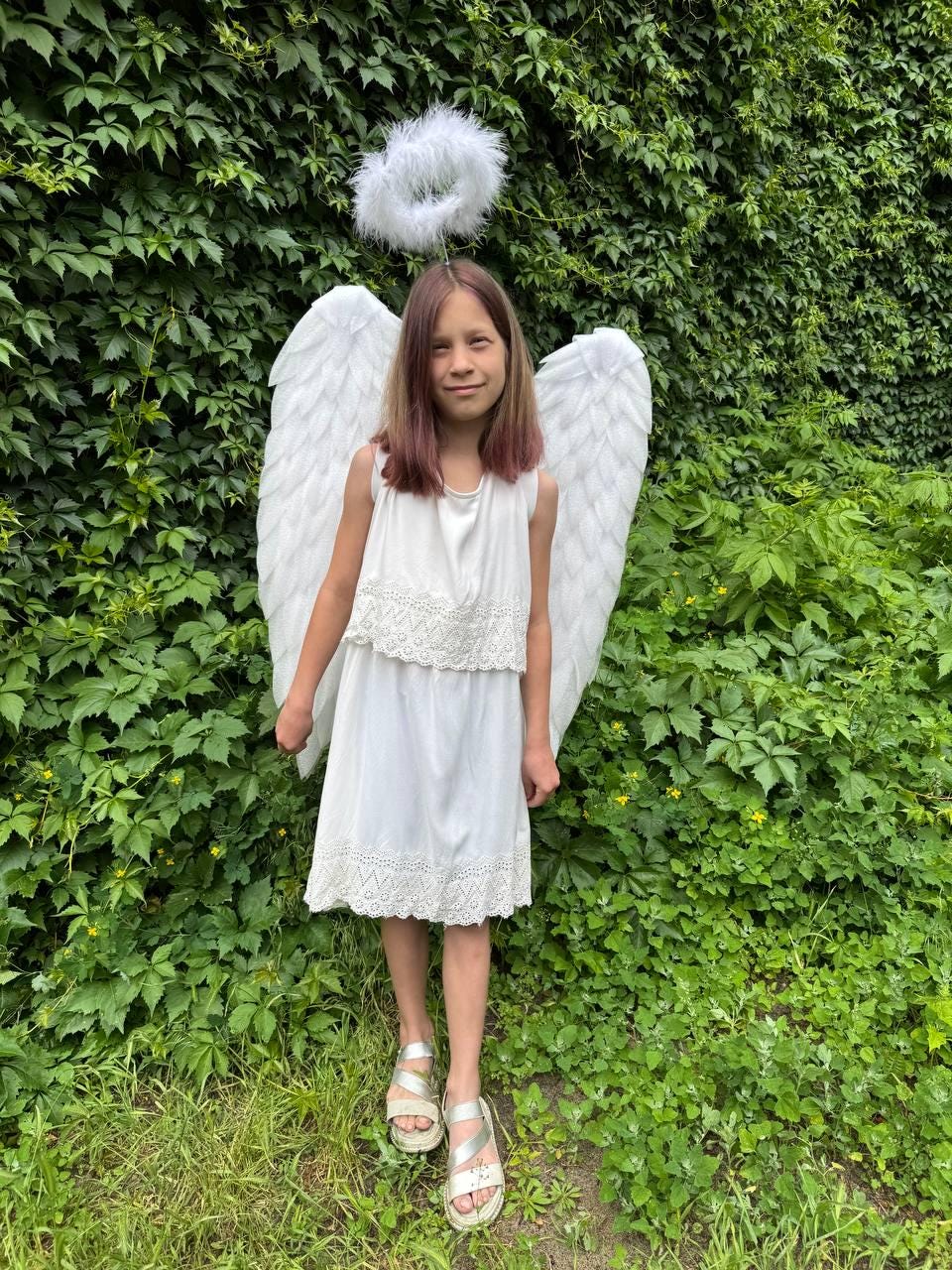 May include: A young person wearing a white dress, silver sandals, and angel wings. A white halo is above the head. The background is a wall of green leaves and grass.