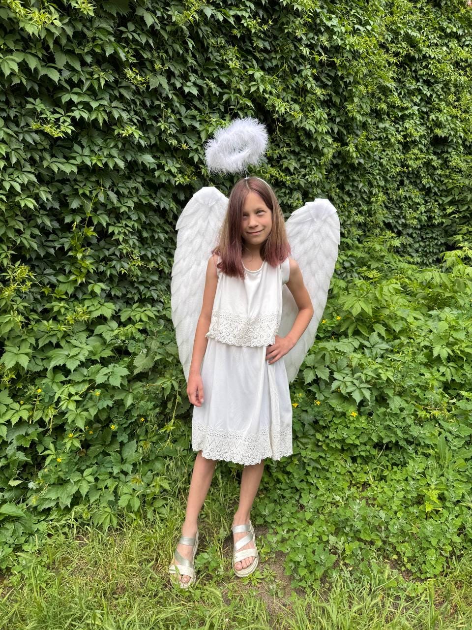 May include: A young person dressed as an angel, wearing a white dress with lace trim, white feathered wings, and a halo. The background is a lush green wall of foliage, with grass in the foreground. The angel is smiling.