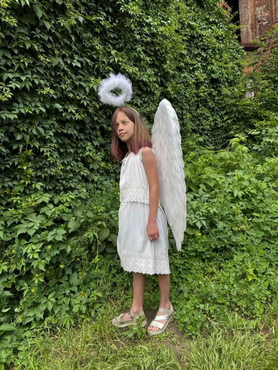 May include: A person wearing a white dress, sandals, and angel wings stands in front of a green leafy background. A white halo is worn on the head. The dress has a lace trim.