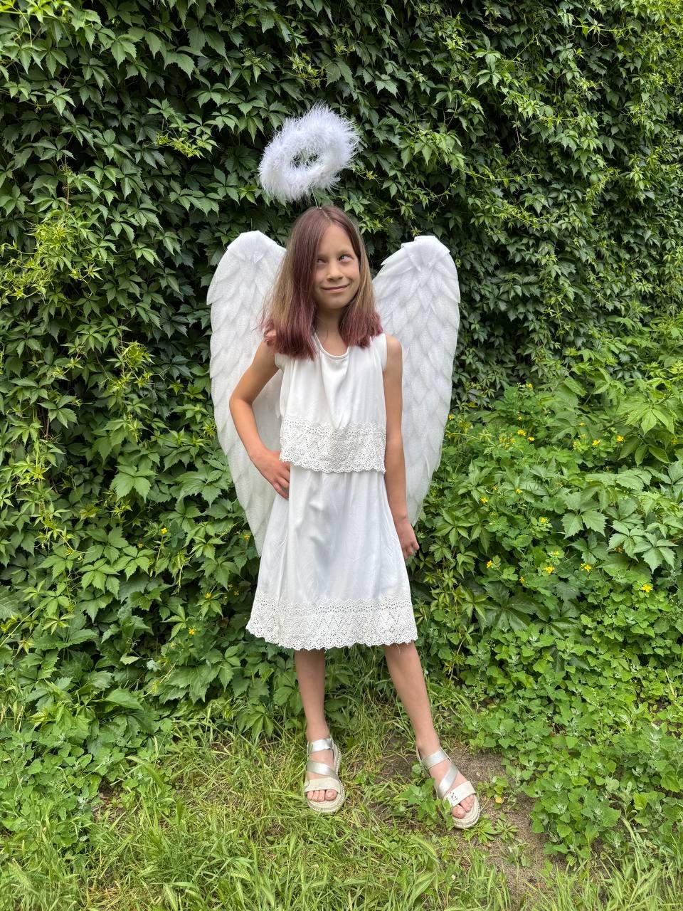 May include: A young person in a white angel costume, including a sleeveless dress with lace trim, large white feathered wings, and a halo. The person is standing in front of a green leafy background, wearing silver sandals.