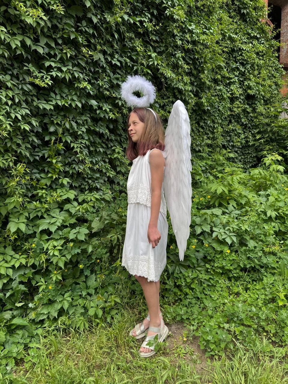 May include: A white angel costume, including a dress with lace trim, feathered wings, and a halo. The costume is worn outdoors against a backdrop of green foliage and grass. The angel costume is suitable for a child.
