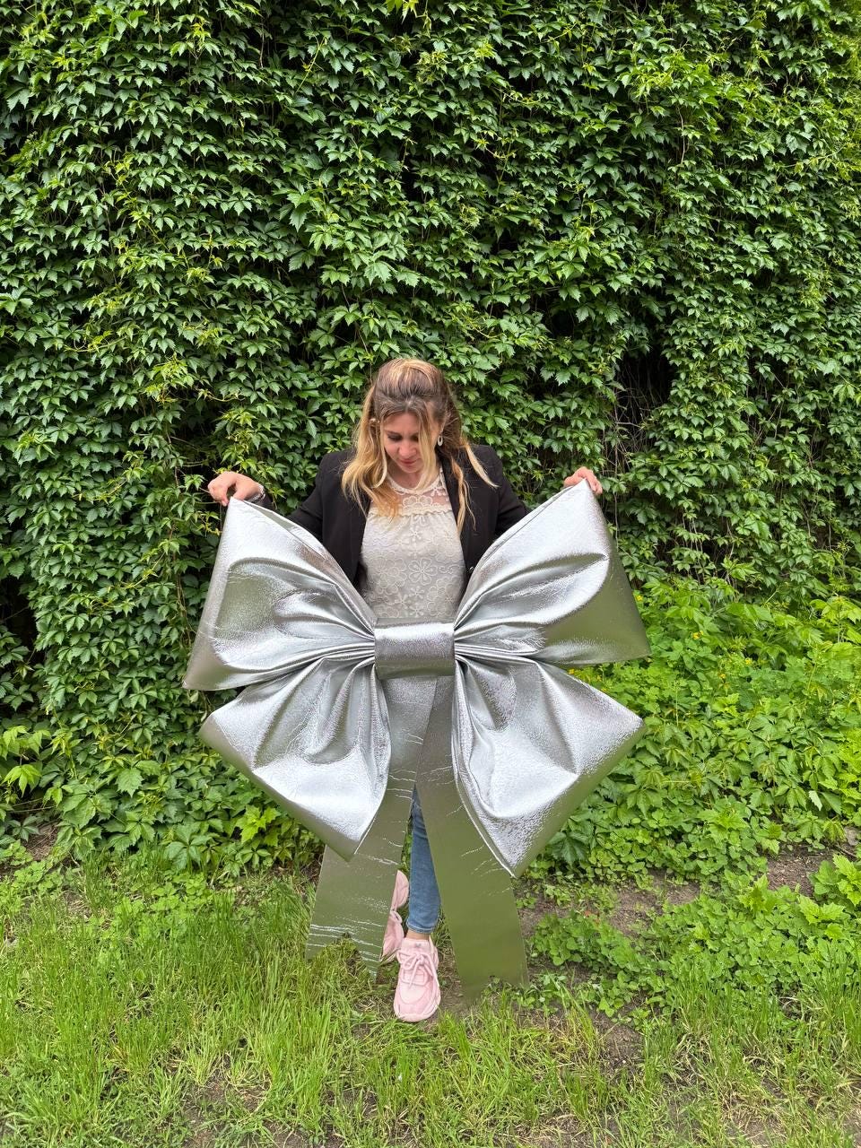 May include: A large, silver metallic bow with a shiny finish. The bow is displayed outdoors against a backdrop of green foliage and grass. The bow is a decorative item, possibly for a party or event.
