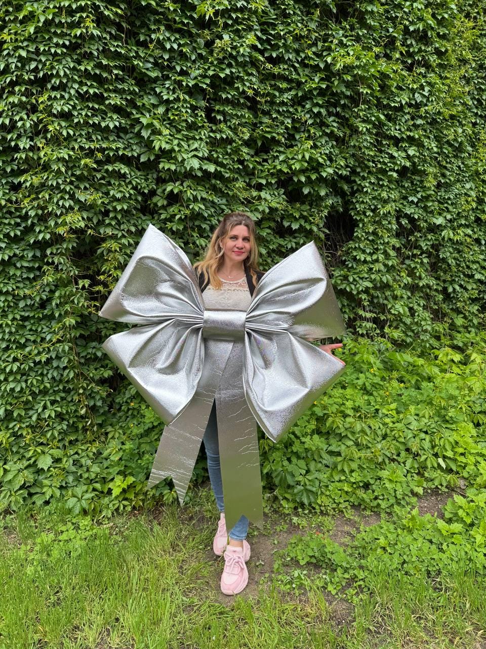 May include: A large, shiny silver bow. The bow is made of a metallic material and has long, flowing ribbons. The bow is large enough to be worn. The background is a wall of green leaves and grass.
