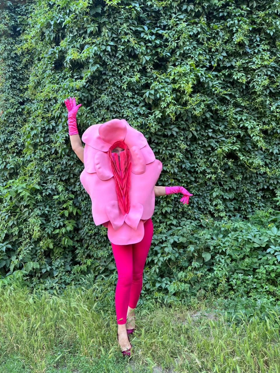 May include: A person wearing a pink costume resembling a flower, with matching leggings and gloves. The costume has a large, open center and is set against a backdrop of green foliage and grass.