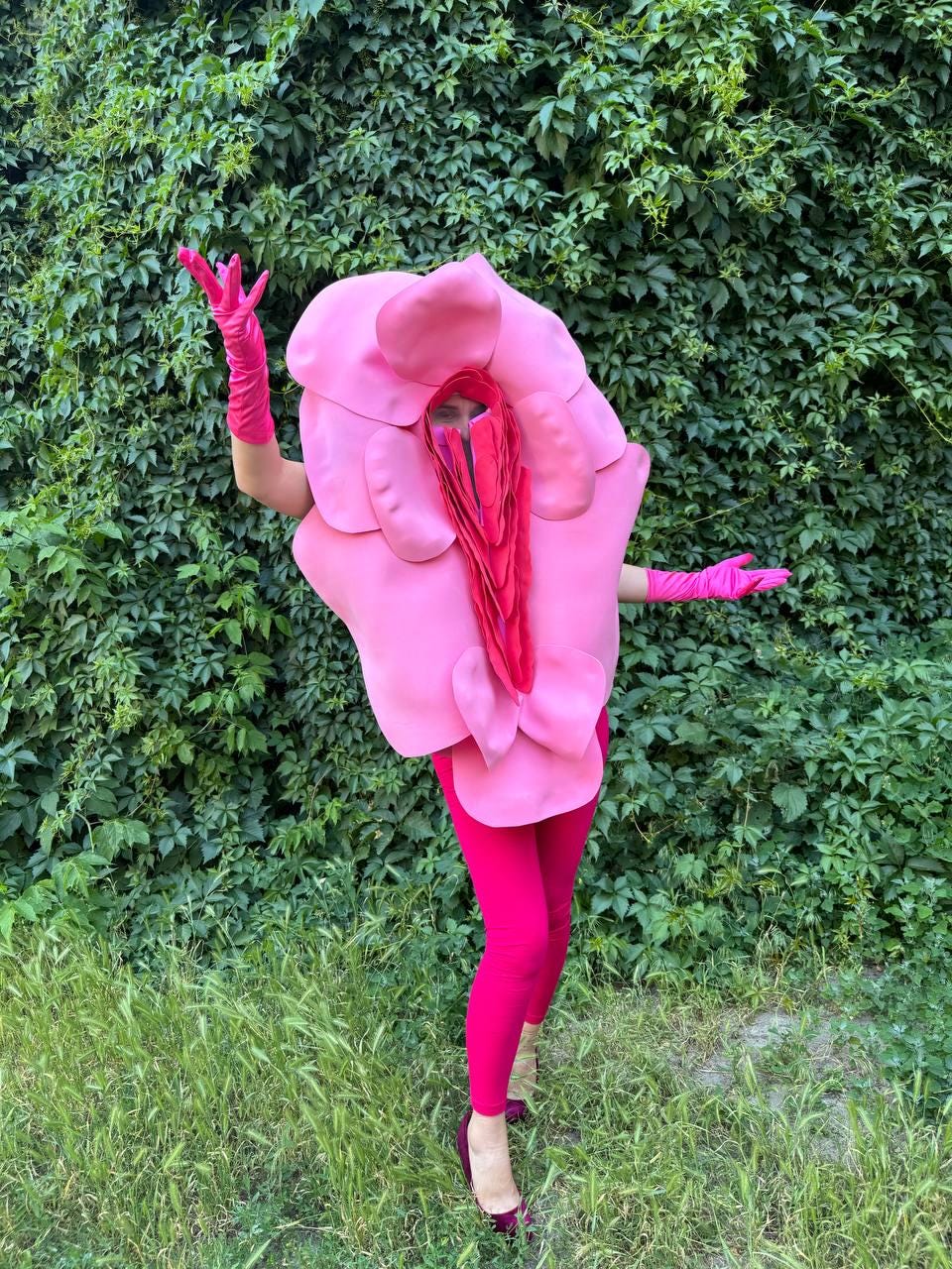 May include: A pink costume resembling a flower, with a large, petal-like structure around the head and shoulders. The costume is paired with pink gloves and leggings. The background is a wall of green foliage.