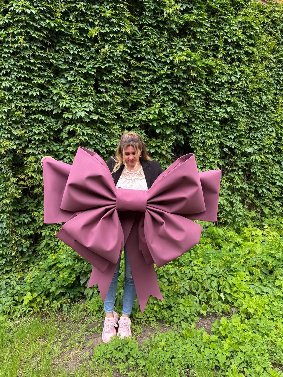 May include: A large, burgundy-colored bow with wide, flowing ribbons. The bow is held in front of a person standing in front of a wall of green foliage. The bow's size suggests it is for decoration or a large gift.