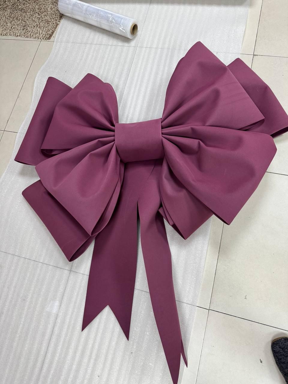 May include: A large, plum-colored decorative bow made of fabric. The bow features multiple loops and long, pointed tails. It is likely used for gift wrapping or event decorations.