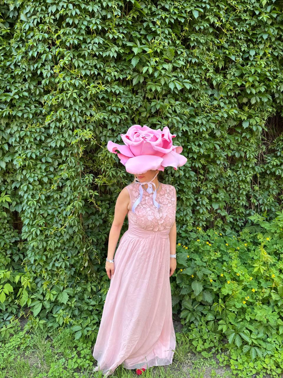 May include: A person wearing a large pink rose headpiece and a light pink dress. The dress has a lace bodice and a flowing skirt. The background is a wall of green leaves and foliage.