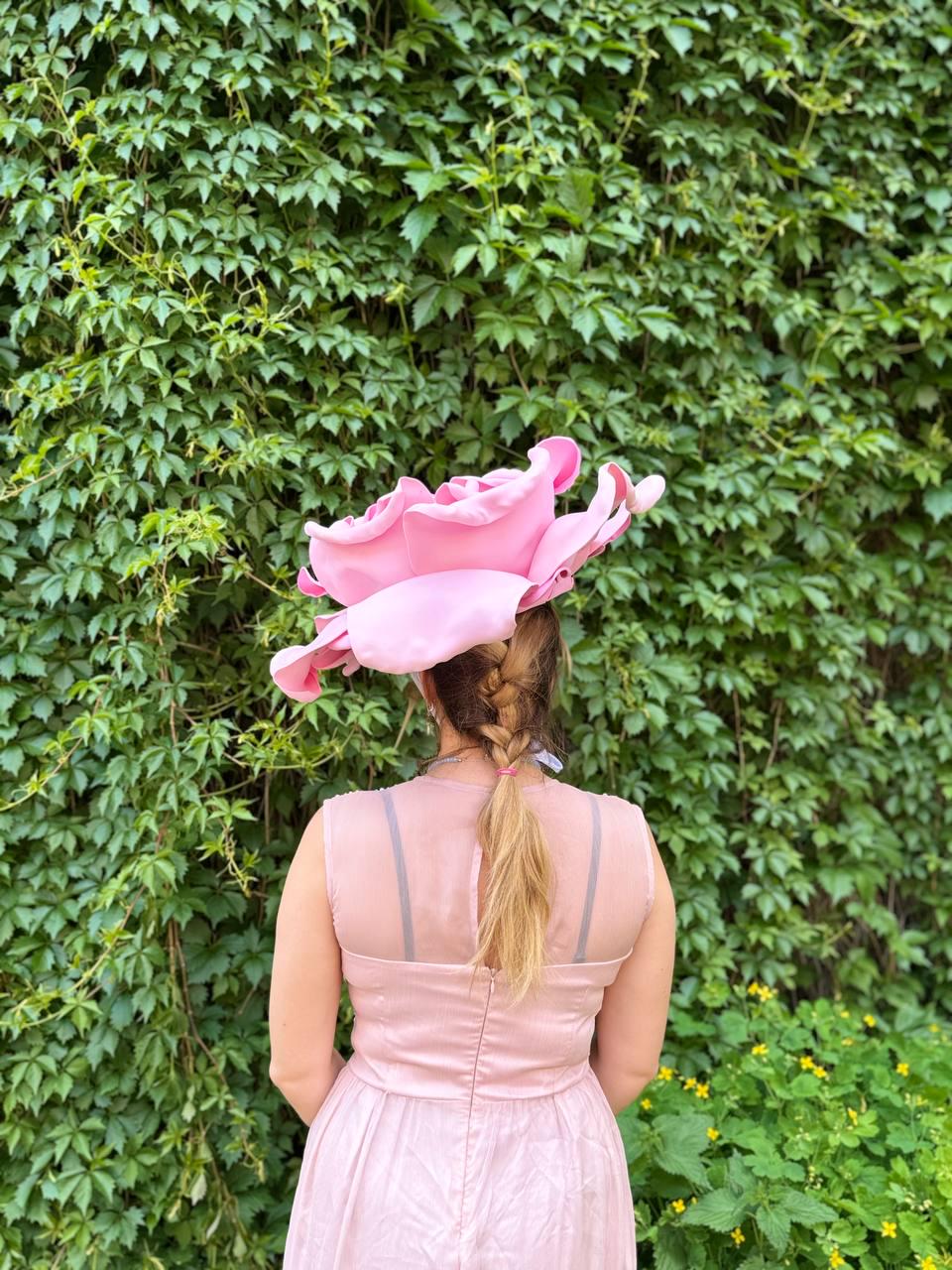May include: A pink dress and a large pink rose-shaped hat are featured in this image. The dress has a fitted bodice and a flowing skirt. The hat is the focal point, with its oversized, petal-like design. The background is a wall of green foliage.