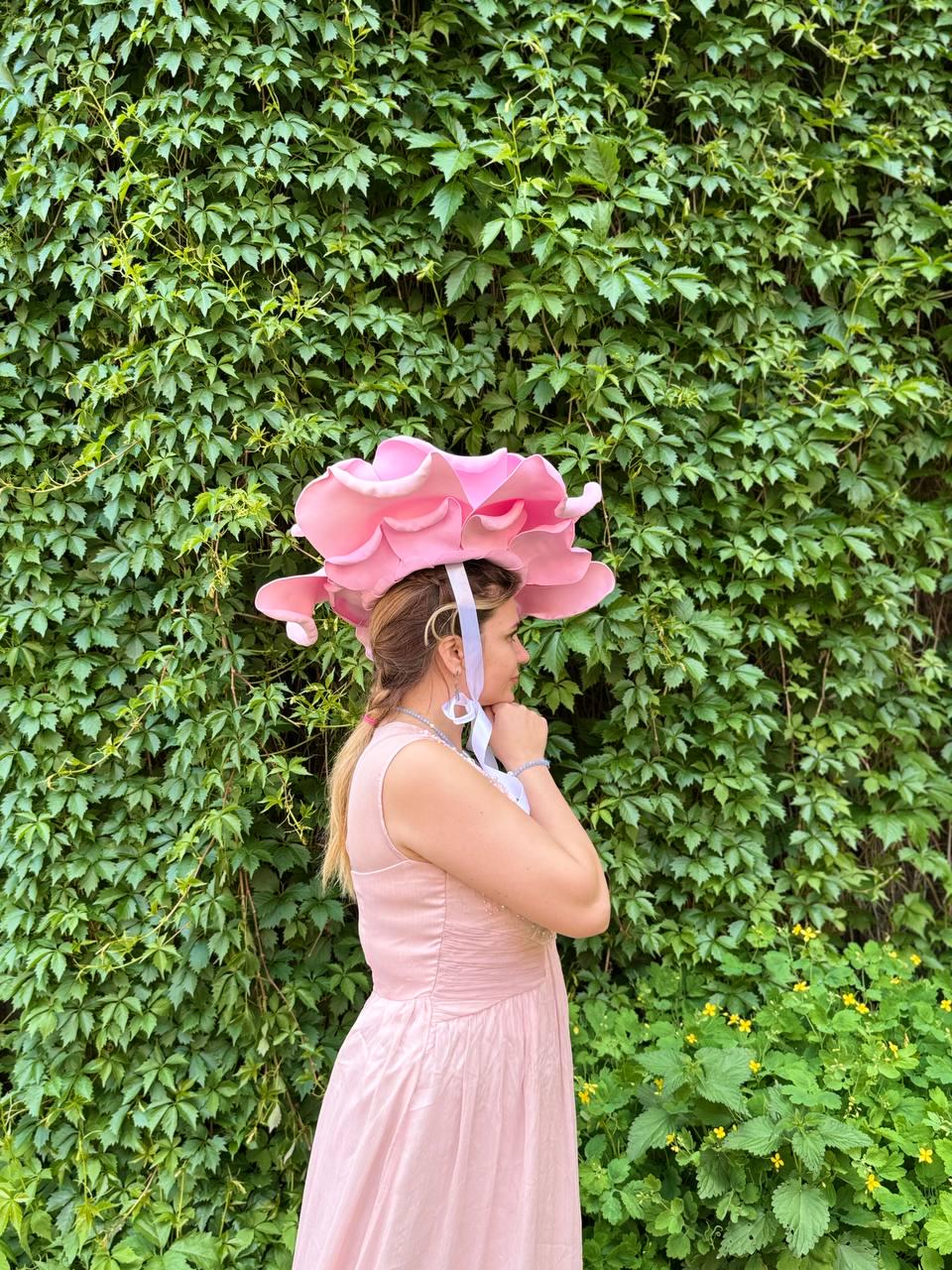 May include: A light pink, flower-shaped hat with multiple petals. The hat is worn with a light pink dress. The background is a wall of green leaves and foliage. The hat is secured with a white ribbon.