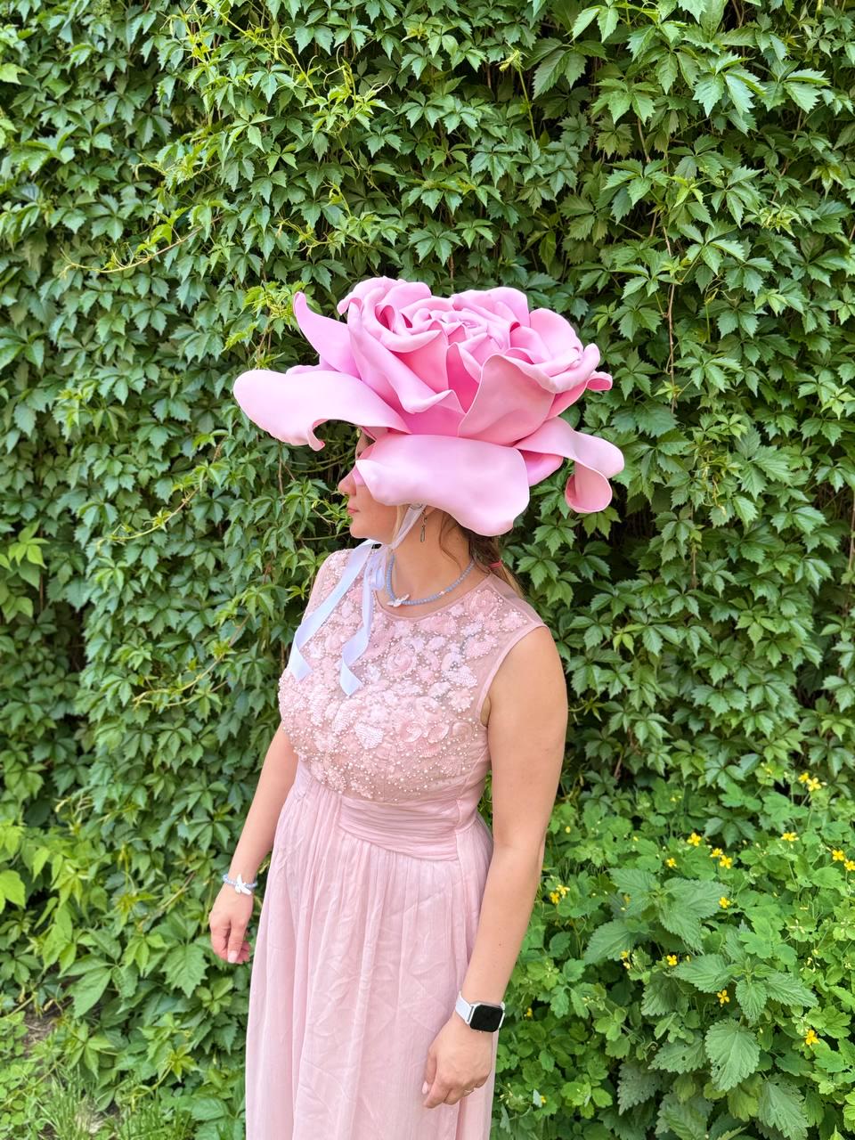 May include: A large, pink rose-shaped hat. The hat is the focal point, with a detailed design mimicking rose petals. The wearer is dressed in a pink dress, with a matching bodice. The background is a lush green wall of foliage.