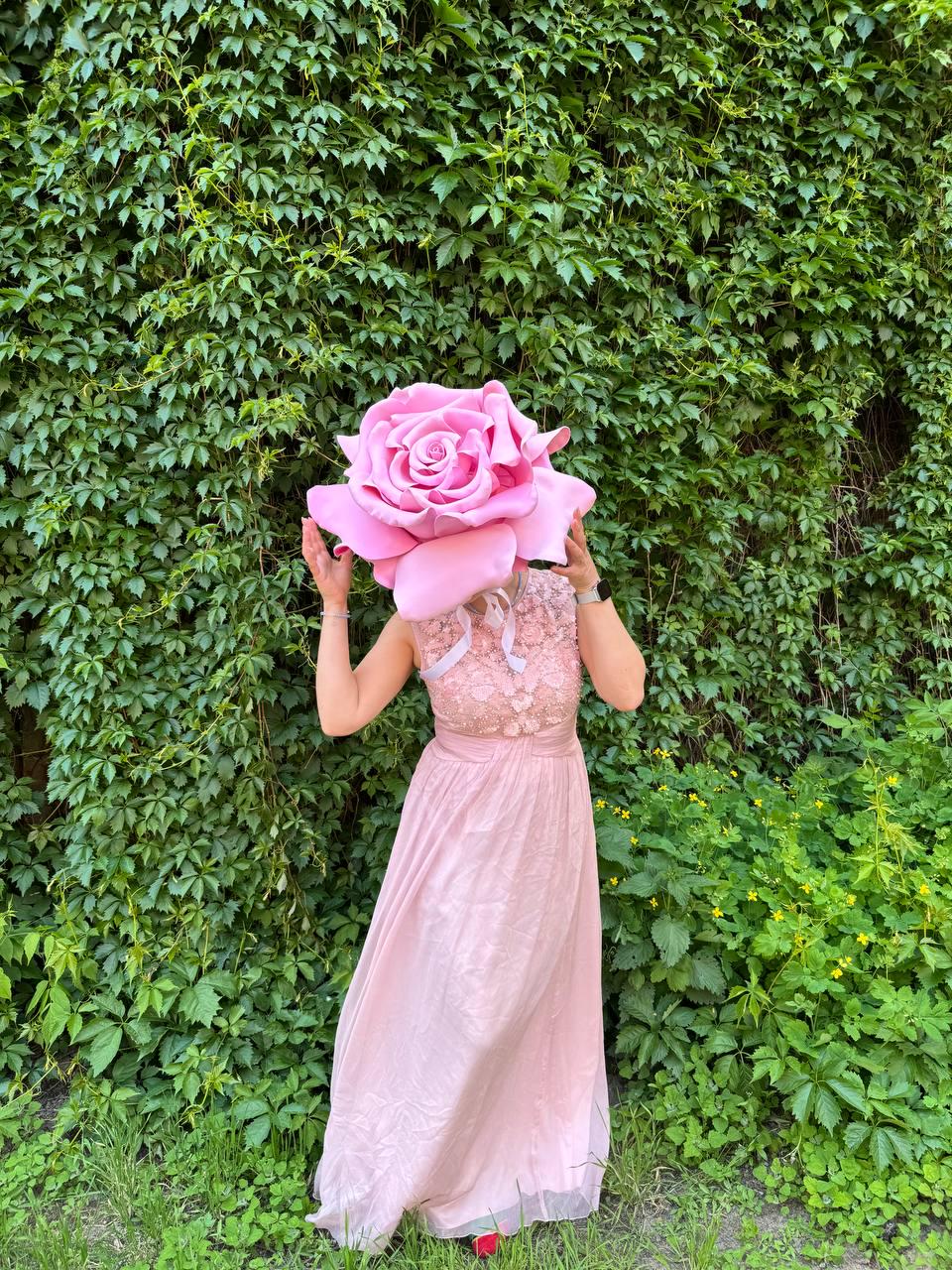 May include: A person wearing a large, pink rose headpiece and a light pink dress. The dress has a lace bodice and a flowing skirt. The background is a wall of green ivy and grass.
