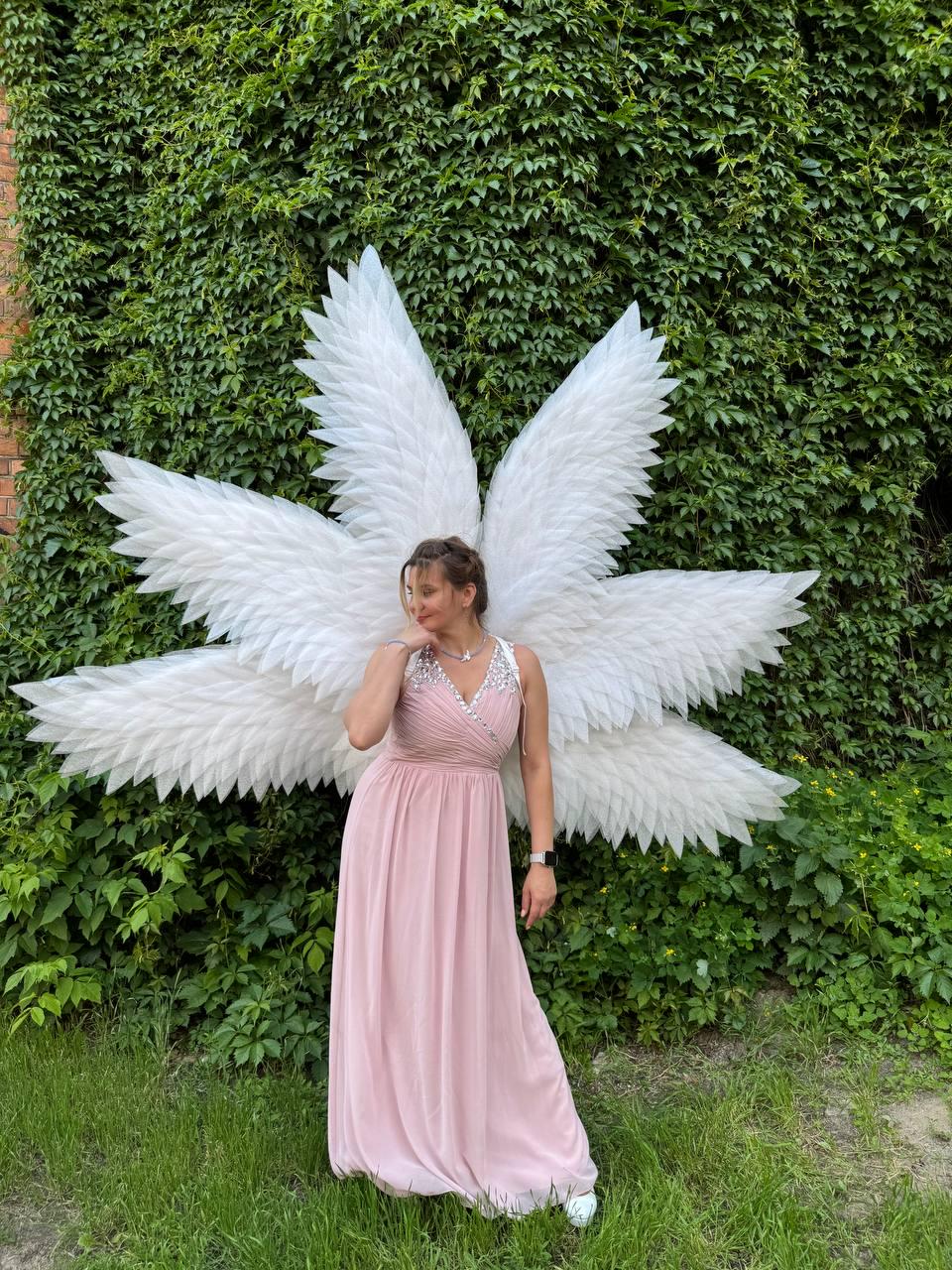 May include: A woman wearing a light pink, floor-length dress and large white angel wings. The wings are made of layered feathers. The background is a wall of green ivy and grass.