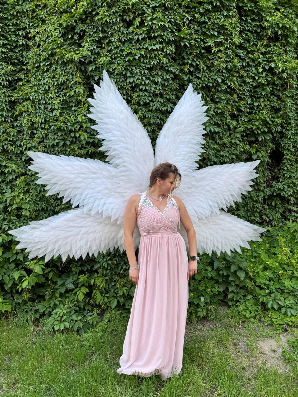 May include: A woman wearing a light pink dress and large white angel wings stands in front of a green ivy wall. The wings are made of white feathers and are spread out behind the woman. The dress has a halter neckline with silver embellishments.