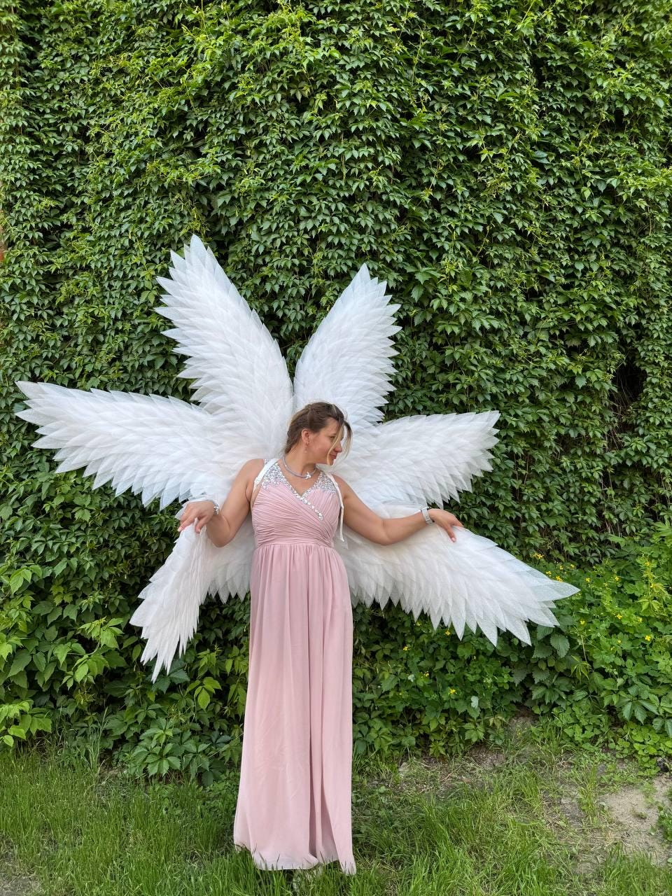 May include: A woman wearing a long, light pink dress and large white angel wings stands in front of a wall of green foliage. The wings are made of white feathers and are spread out behind her. The dress has a V-neckline and a flowing skirt.