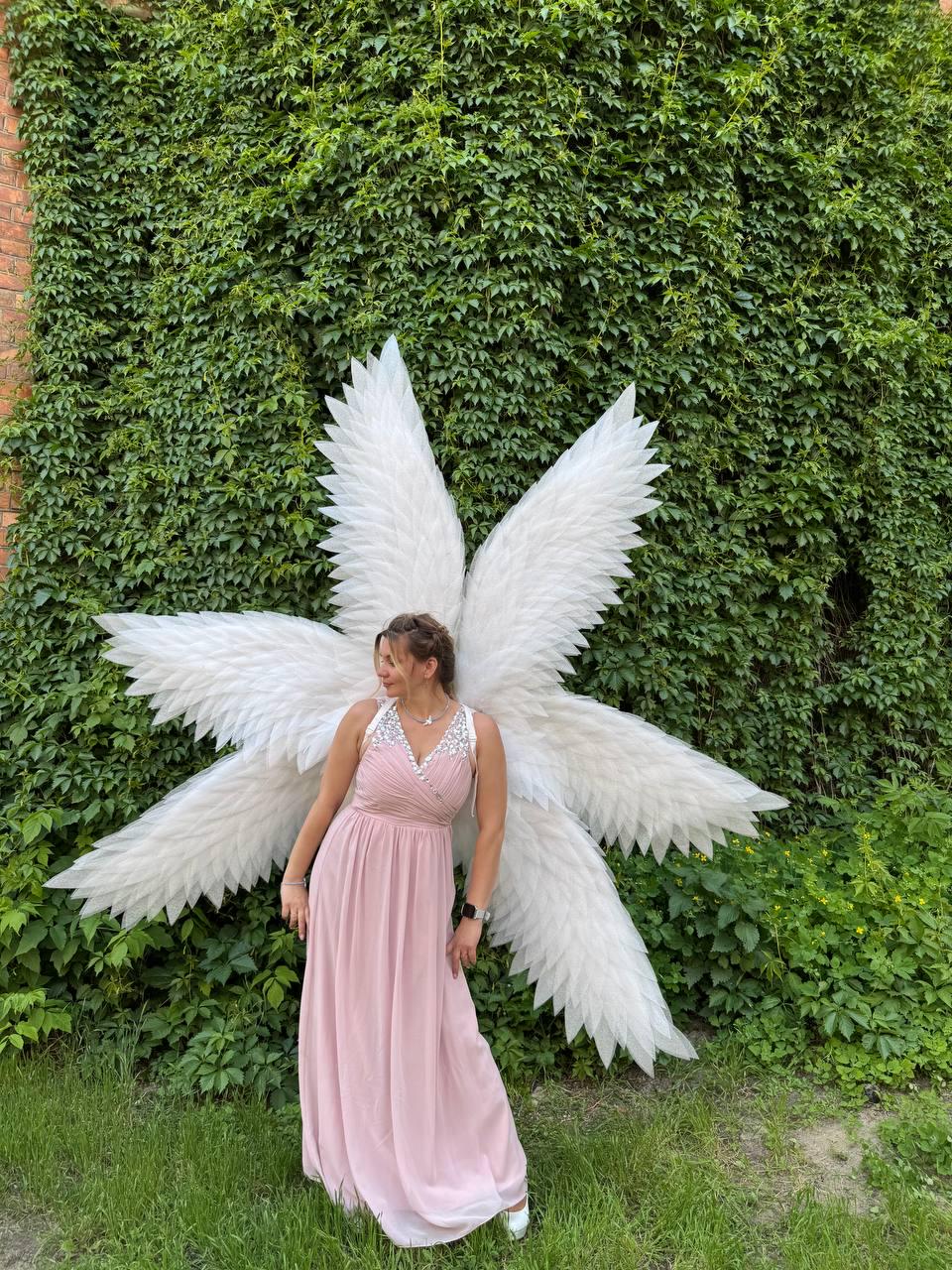 May include: A woman wearing a light pink dress and large white angel wings stands in front of a wall covered in green foliage. The wings are made of white feathers and are spread out behind her.
