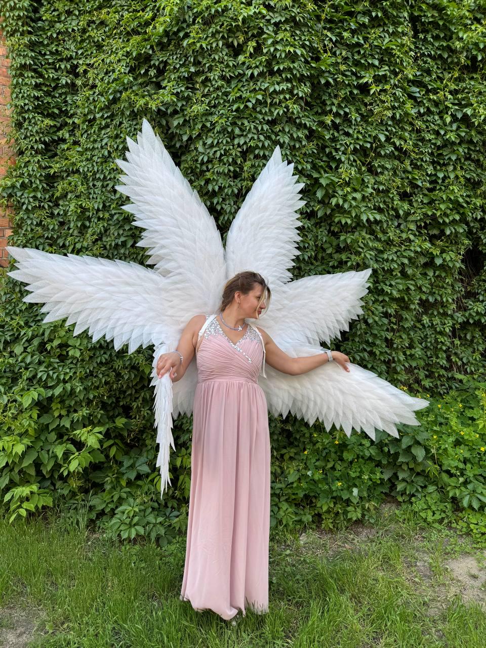 May include: A woman wearing a long, light pink dress and large white angel wings stands in front of a wall covered in green foliage. The wings are made of white feathers and are attached to the back of the dress.
