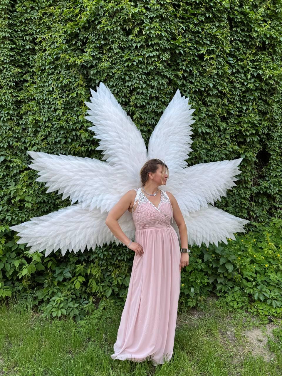 May include: A person wearing a light pink dress and large white angel wings stands in front of a wall of green foliage. The wings are made of white feathers and are spread out behind the person. The dress has a beaded neckline.