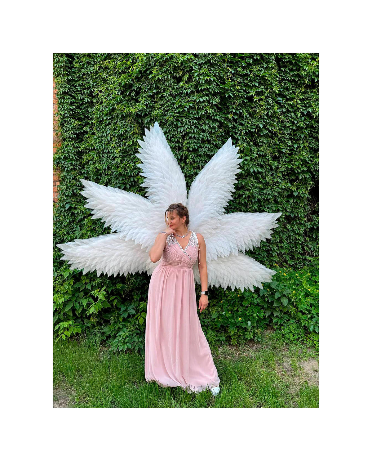 May include: A woman wearing a long, light pink dress and large white angel wings stands in front of a wall covered in green foliage. The dress has a V-neckline with white lace trim. The wings are large and feathery.