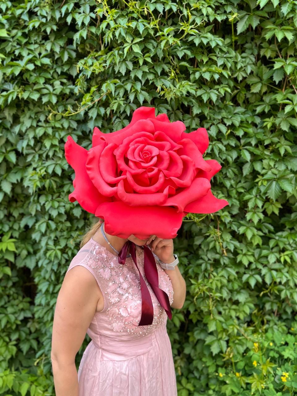 May include: A large, vibrant red rose headpiece, obscuring the face of a person wearing a pink dress with floral embellishments. A burgundy ribbon is tied around the neck. The background is a dense wall of green foliage.