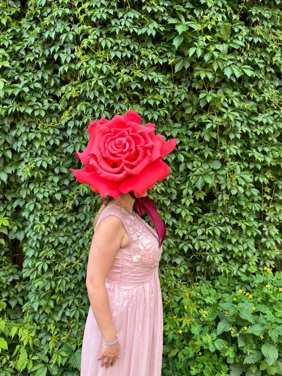 May include: A woman wearing a large, vibrant red rose headpiece and a pale pink dress. The dress has a beaded bodice and a flowing skirt. The background is a lush green wall of ivy.