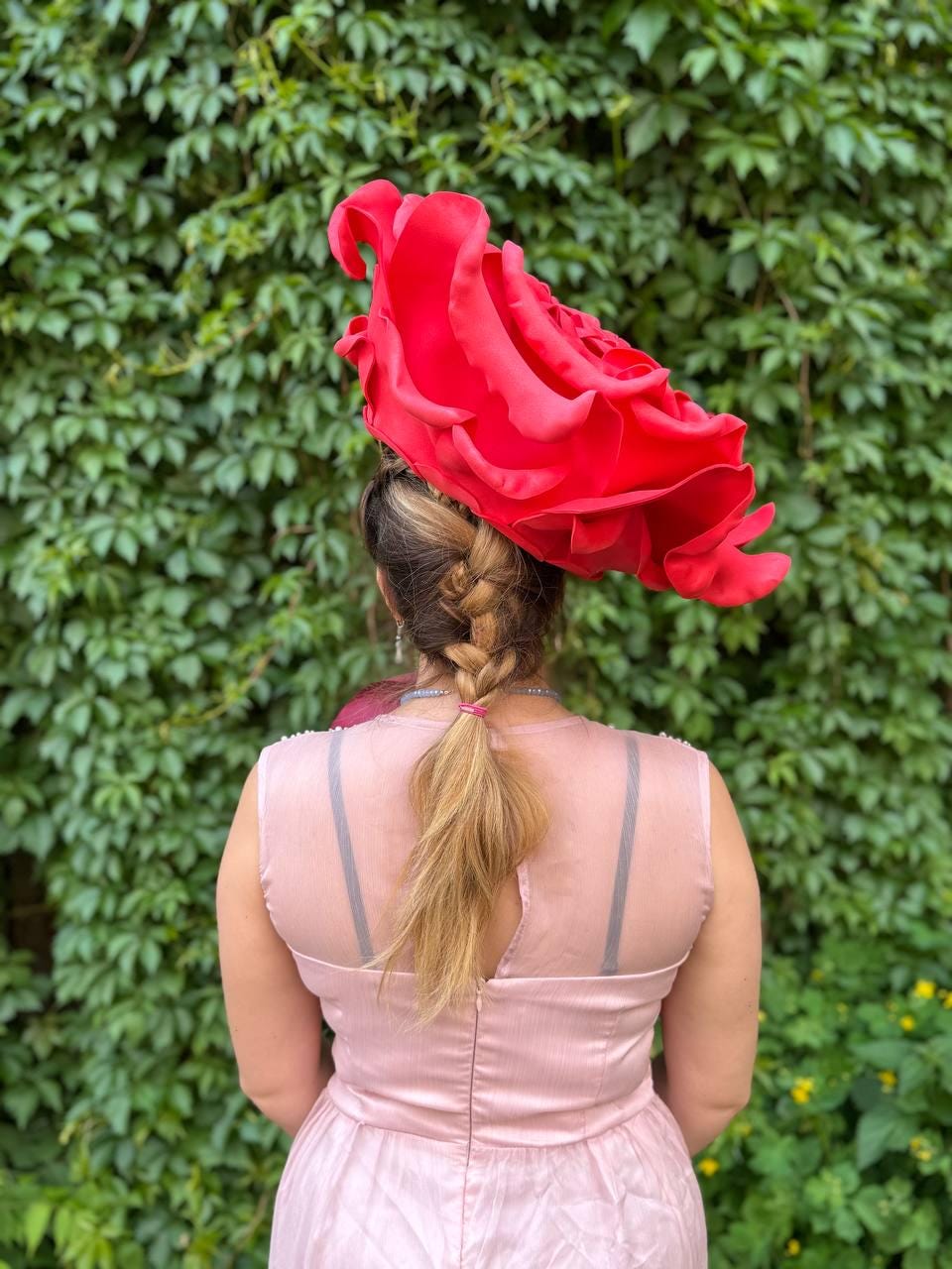 May include: A large, vibrant red hat shaped like a rose, worn by a person in a light pink dress. The dress has a sheer back with visible straps. The person's hair is styled in a braid. The background is a wall of green leaves.