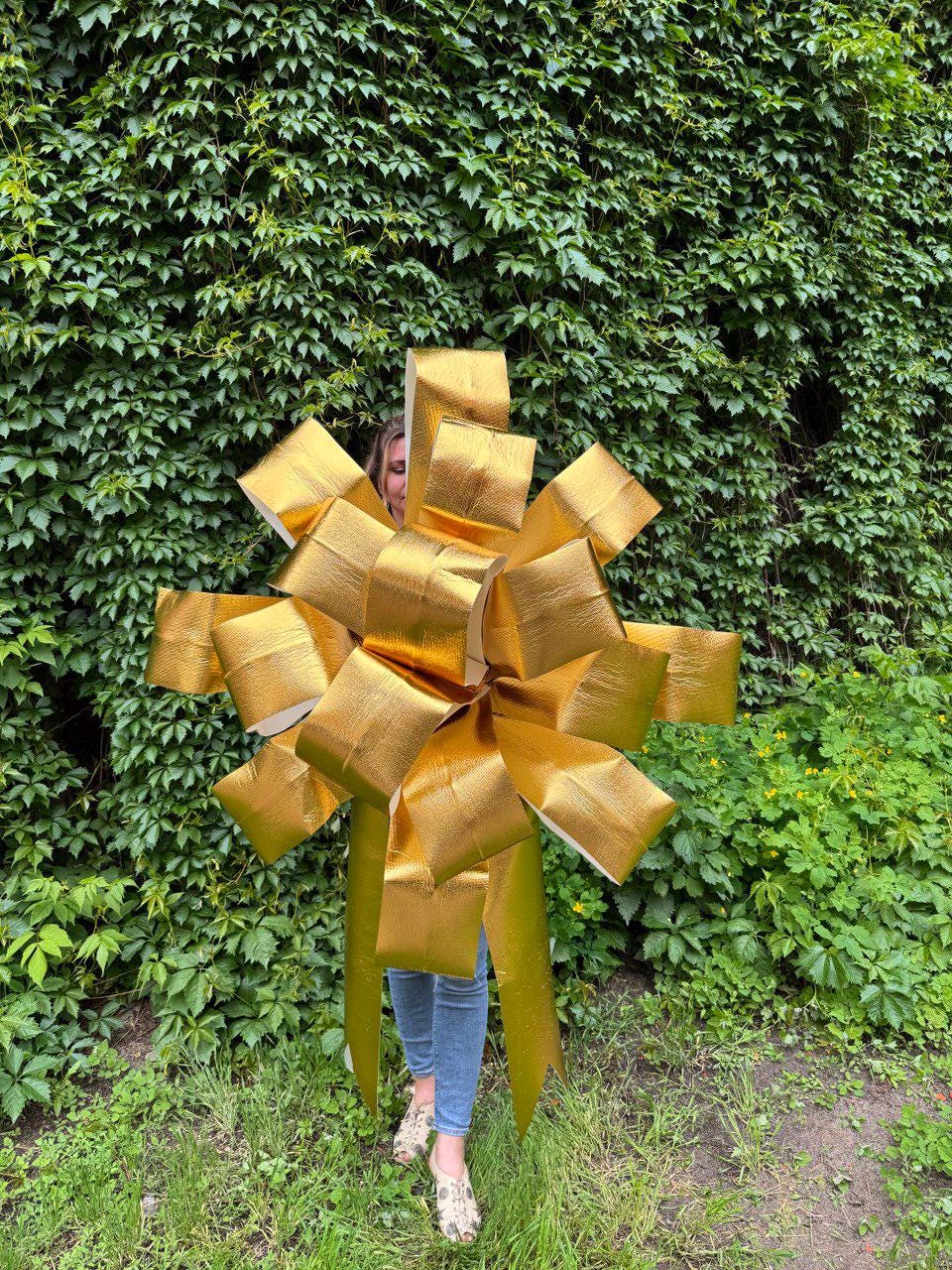 May include: A large, oversized decorative bow made of metallic gold material with a textured finish. The bow features multiple layered loops and two long, pointed tails extending downward, creating a voluminous and star-like shape.