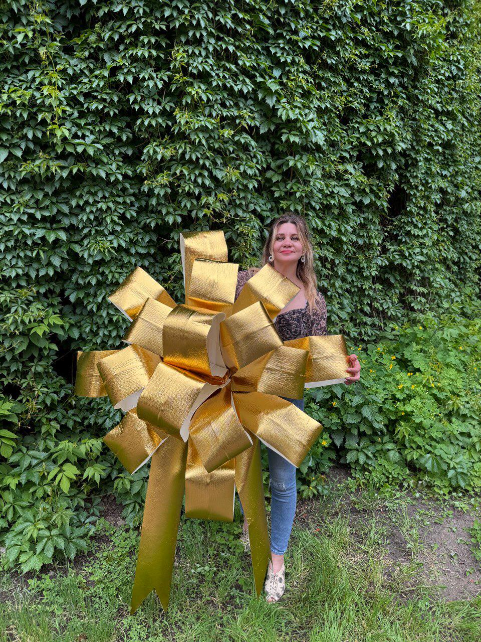 May include: A large, oversized decorative bow with a metallic gold finish and a textured surface. The bow features multiple wide, structured loops and two long, pointed tails extending downward, creating a voluminous and eye-catching display piece.