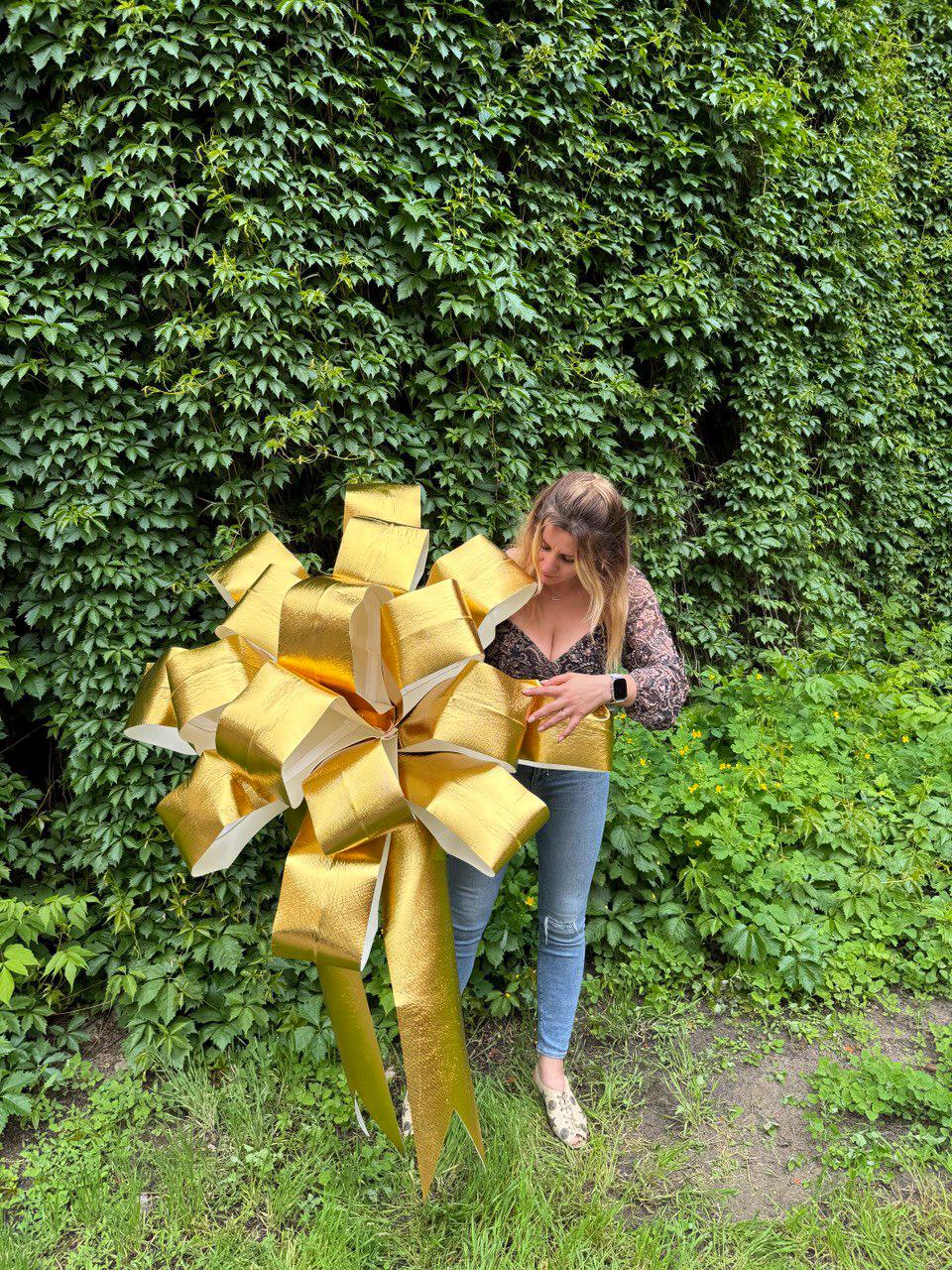 May include: A large, oversized decorative bow with a metallic gold finish and a textured surface. The bow features multiple voluminous loops and two long, pointed ribbon tails extending downward, creating a full and rounded structural shape.