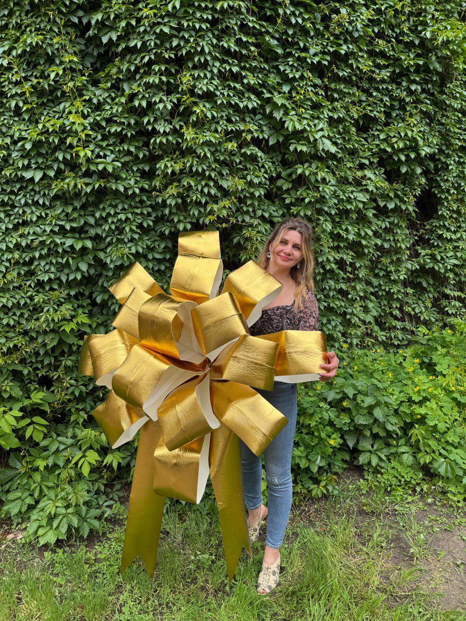 May include: A large, oversized decorative bow with a metallic gold finish on the front and a white underside. The bow features multiple layered loops and two long, pointed tails extending downward, creating a voluminous and structured appearance.