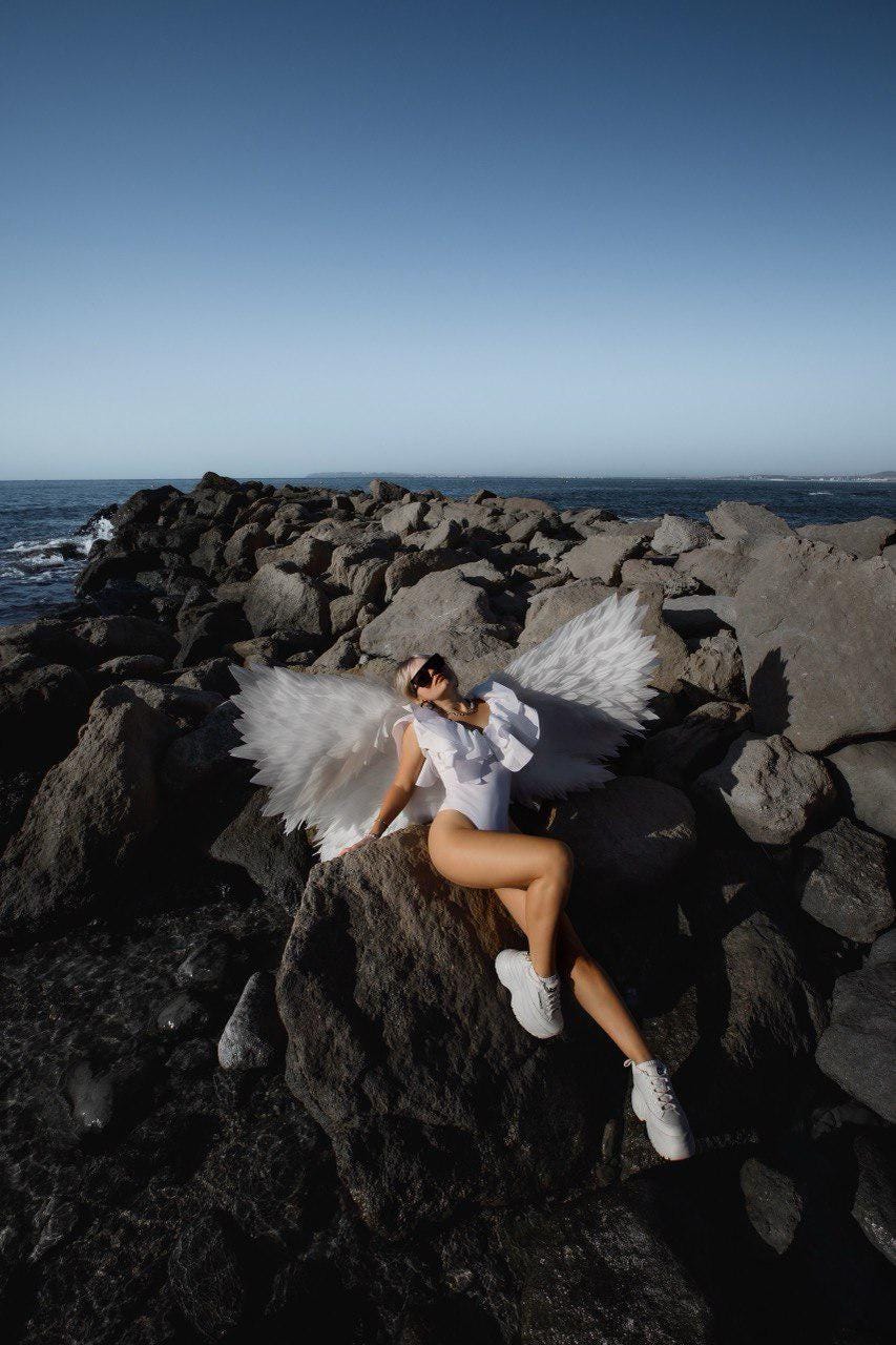 May include: A person wearing a white bodysuit, sunglasses, and white sneakers, posing with large white angel wings on a rocky shoreline. The background features the ocean and a clear blue sky.