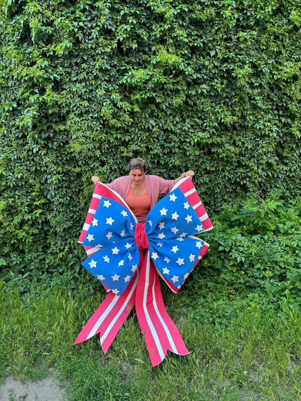 May include: A large patriotic bow with a blue center adorned with white stars and red and white striped ribbons. The bow is displayed against a backdrop of lush green foliage and grass.