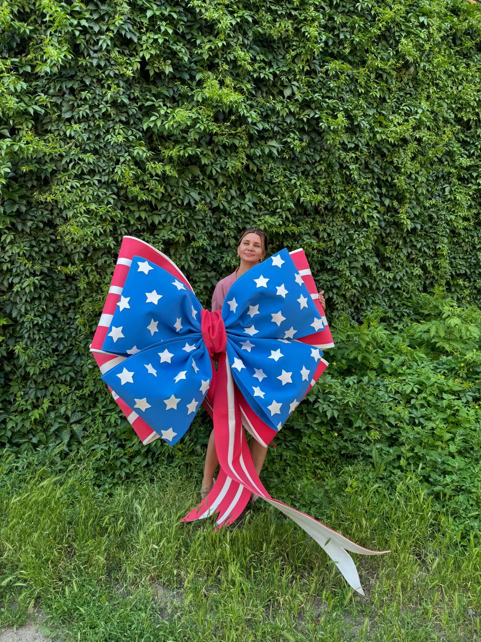 May include: A large decorative bow in patriotic colors. The bow features a blue center with white stars, red and white striped edges, and long red and white striped streamers. The bow is held in front of a green leafy background.