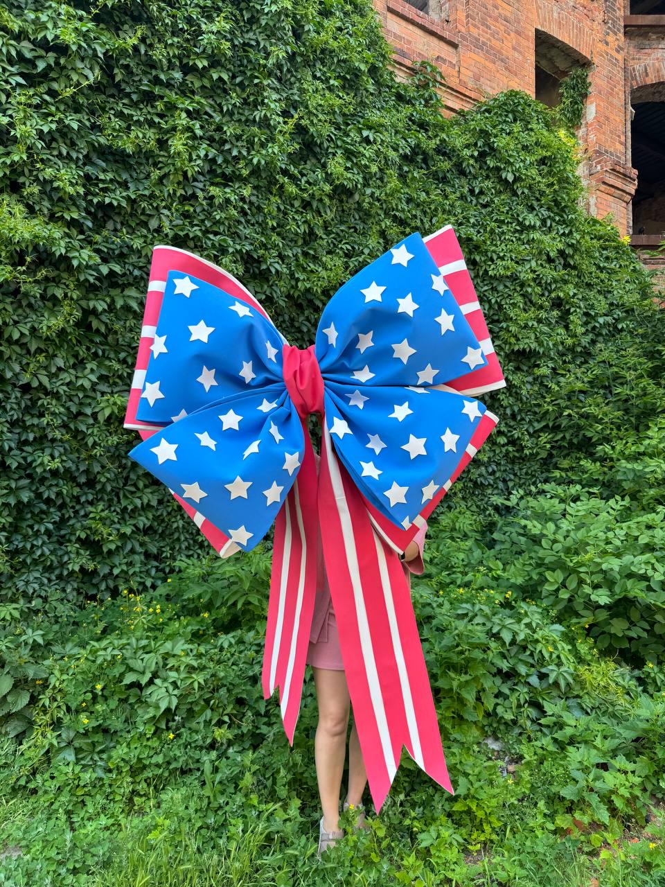 May include: A large decorative bow featuring a patriotic design. The bow has a blue center adorned with white stars, red and white striped edges, and long red and white striped streamers. The bow is held in front of a person standing in front of a green leafy background.