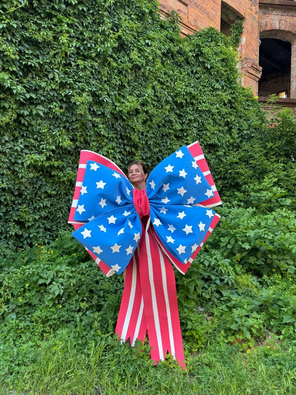 May include: A large decorative bow with a patriotic design. The bow features blue sections with white stars, red and white striped ribbons, and a red center. The bow is displayed against a backdrop of green foliage and a brick building.