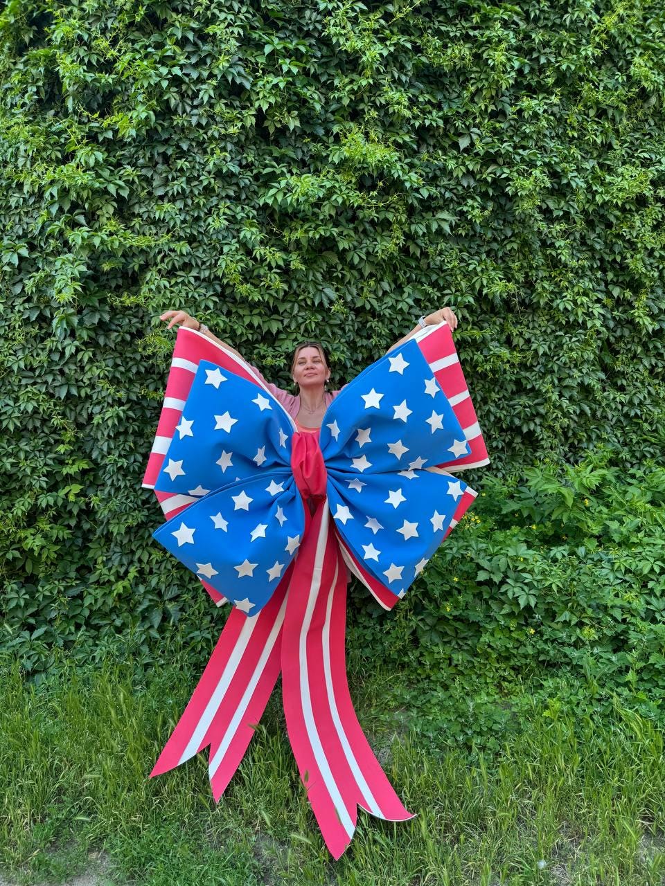 May include: A large decorative bow with a patriotic theme. The bow features a blue center with white stars and red and white striped ribbons. The bow is displayed against a backdrop of green foliage.