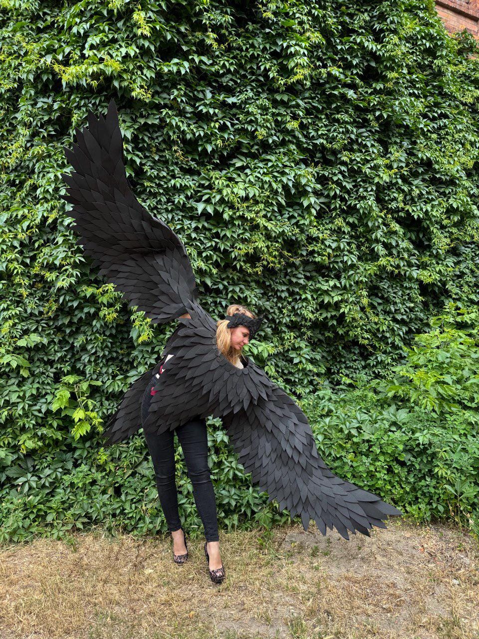 May include: A person wearing large, black, feathered costume wings, standing in front of a wall of green ivy. The wings are spread wide, showcasing their intricate design. The person is wearing black pants and heels.