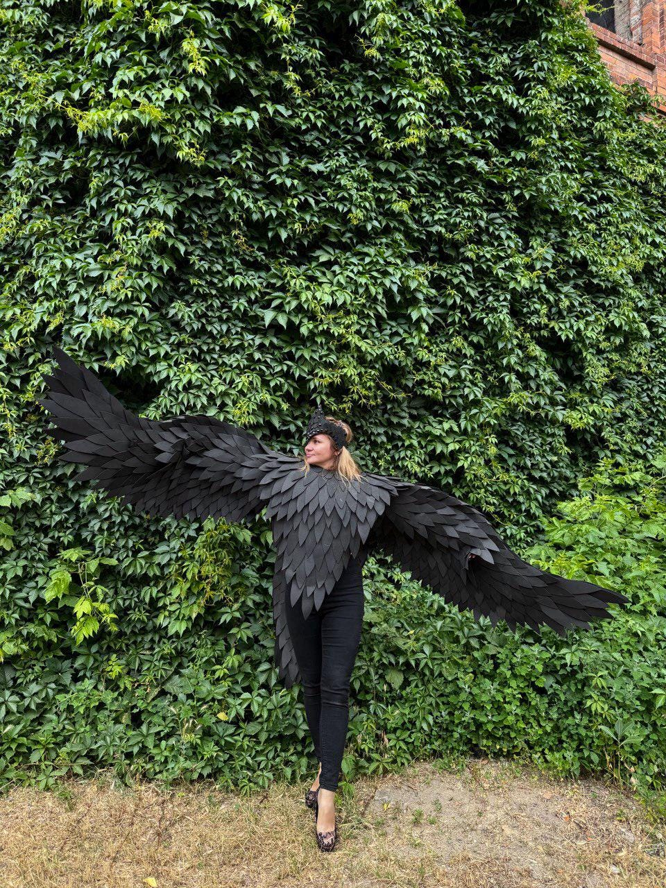 May include: A person wearing a black costume with large, feathered wings, a matching collar, and a headpiece, posing in front of a wall of green foliage. The costume is a striking, theatrical design.