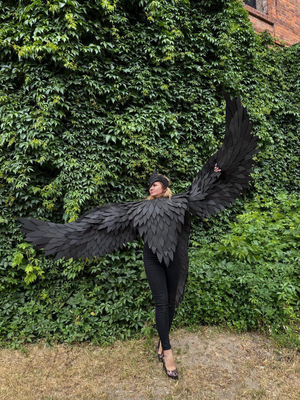 May include: A person wearing a black raven costume with large, layered wings. The costume includes a matching headpiece and is set against a backdrop of lush green foliage and a brick building.