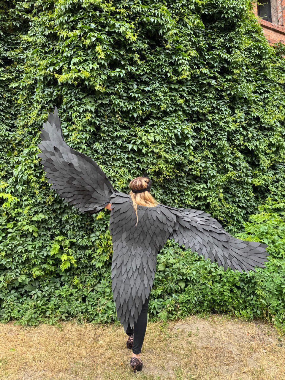 May include: A person wearing large, black, feathered wings, standing in front of a wall of green foliage. The wings are spread wide, showcasing their intricate design and texture. The wings appear to be made of individual, overlapping pieces.