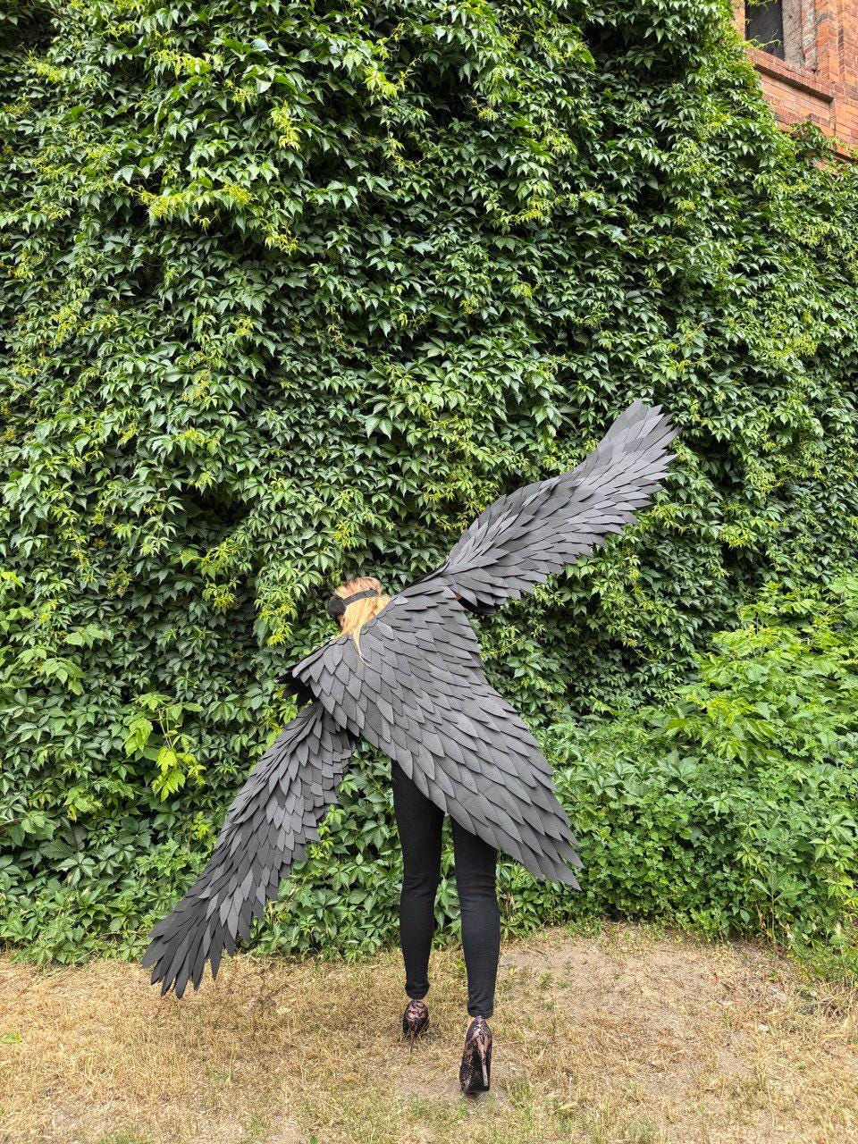May include: A person wearing large, black, feathered wings, standing in front of a wall covered in green ivy. The wings are spread wide, and the person is wearing black pants and patterned heels. The wings appear to be made of individual, overlapping pieces.