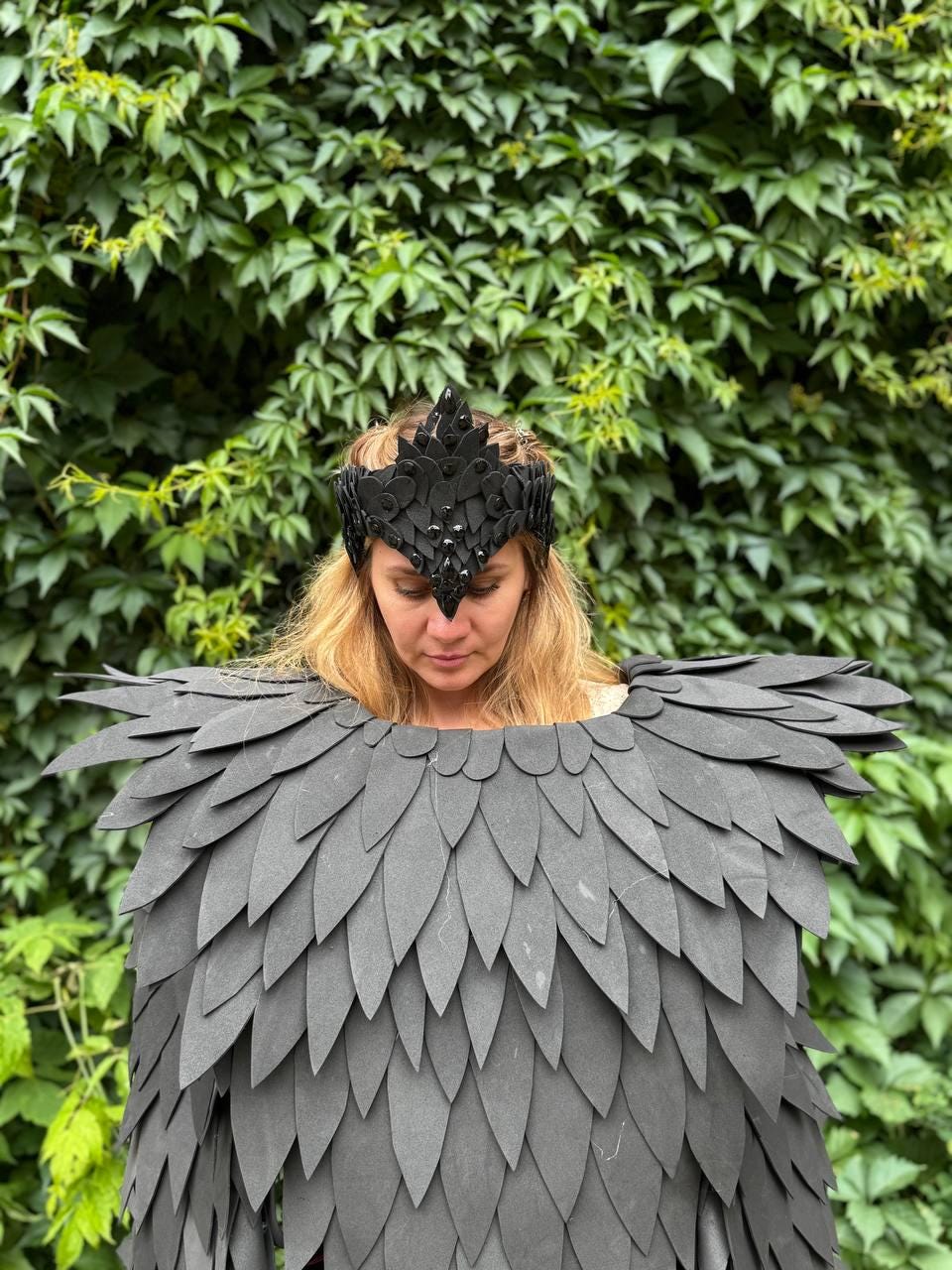 May include: A black crown with a pointed design and a matching black cape with layered feather-like details. The cape covers the shoulders and chest. The background is a wall of green leaves.