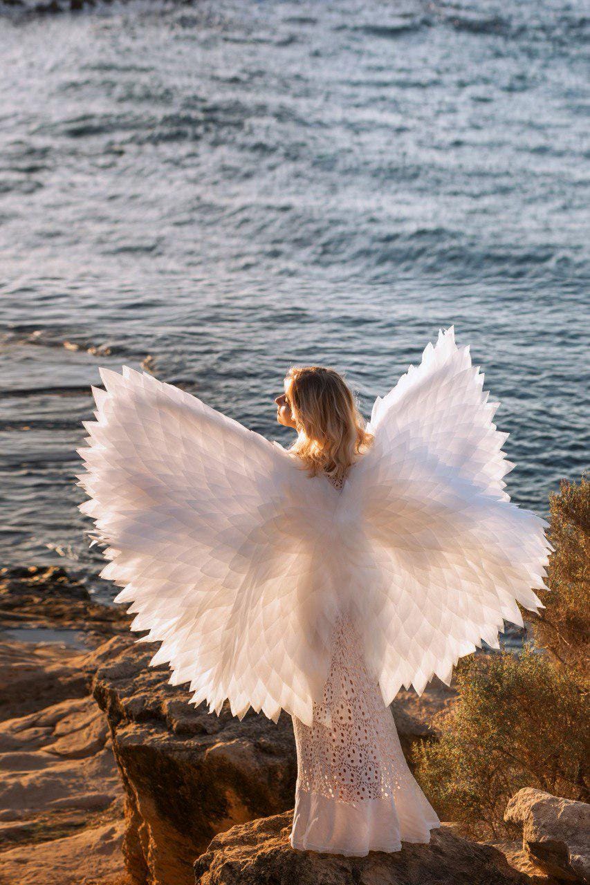 May include: A person wearing large white angel wings stands on a rocky shore, facing the ocean. The wings are detailed and spread wide. The person is wearing a white dress. The scene is bathed in sunlight.