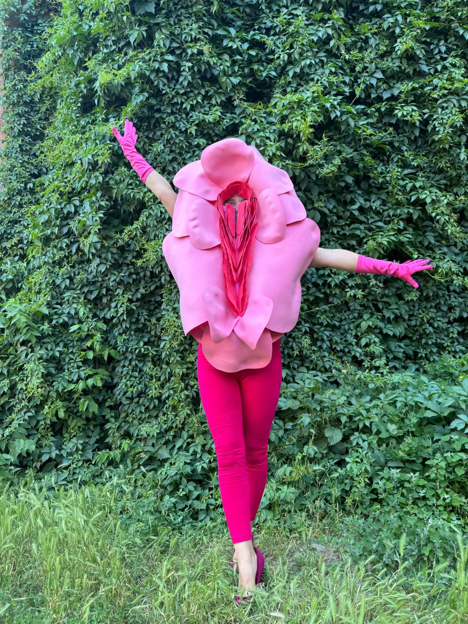 May include: A person wearing a pink costume resembling a flower, with a large pink headpiece and matching gloves. The person is also wearing bright pink leggings and shoes, standing in front of a green leafy background.