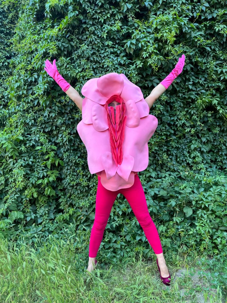 May include: A person wearing a pink flower costume with matching pink leggings, gloves, and shoes, standing in front of a green leafy background. The costume has a large, detailed flower design.